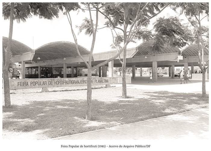   4 J- Feira Popular de Hortifrutis (1981) - Acervo Público do DF Foto de um grande galpão coberto com alambrado claro, em uma mureta com inscritos: Feira Popular de Hortifrutigranjeiro de Planaltina Algumas pessoas entram no galpão, várias árvores sombreiam o local. Realização: Academia Planaltinense de Letras, Artes e Ciências (APLAC), pelo Ecomuseu Pedra Fundamental e pelo Coletivo Nativo Audiodescrição produzida pelo Instituto de Promoção das Pessoas com Deficiência Visual Audiodescritora: Elma Lúcia Rodrigues Consultor: Fernando Rodrigues Este projeto é promovido com recursos do Fundo de Apoio a Cultura do DF